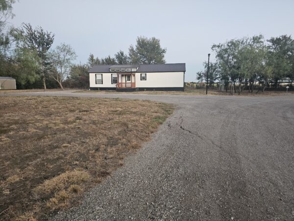 A small, single-story house with a porch sits near a gravel driveway, surrounded by dry grass and trees, under a clear sky.