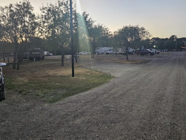 A gravel road runs through an RV park at dusk, with parked cars, RVs, and trees scattered throughout the area under a partly cloudy sky.