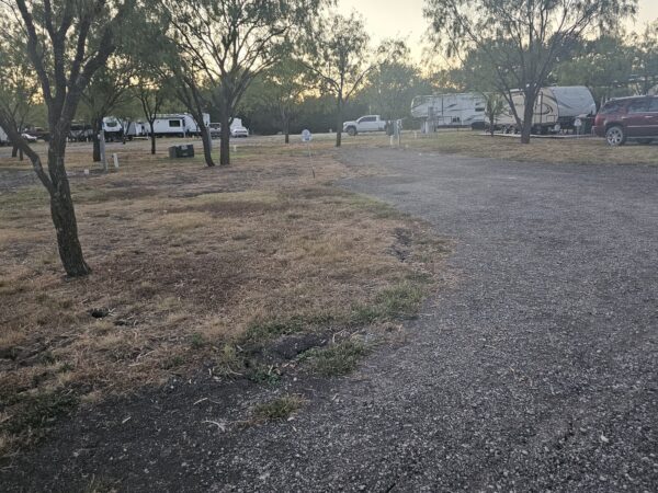 A gravel path curves through a campground with parked RVs, cars, and scattered trees. The grass is dry and patchy, and the sky appears to be at sunset or sunrise.