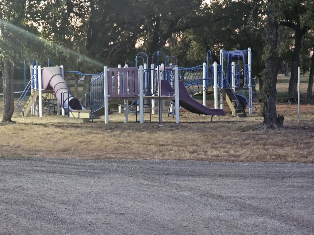 A playground with slides, climbing structures, and bridges sits on dry grass near trees, with a gravel area in the foreground.