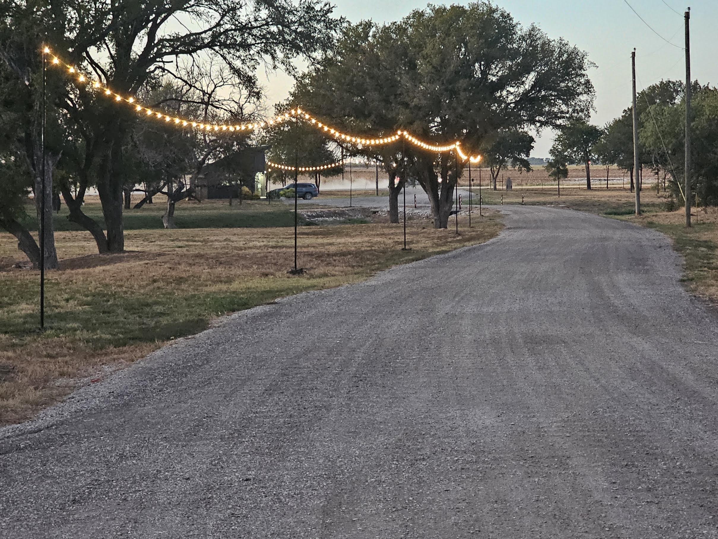 A gravel road curves past trees and grassy areas, with string lights hanging above on poles, creating a festive atmosphere in an open rural setting.