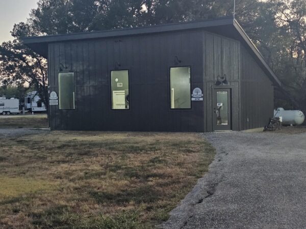 A dark-paneled building with three tall windows, a lit glass door, and outdoor lights. A curved driveway and dry grass are in the foreground. Trees and RVs are visible in the background.