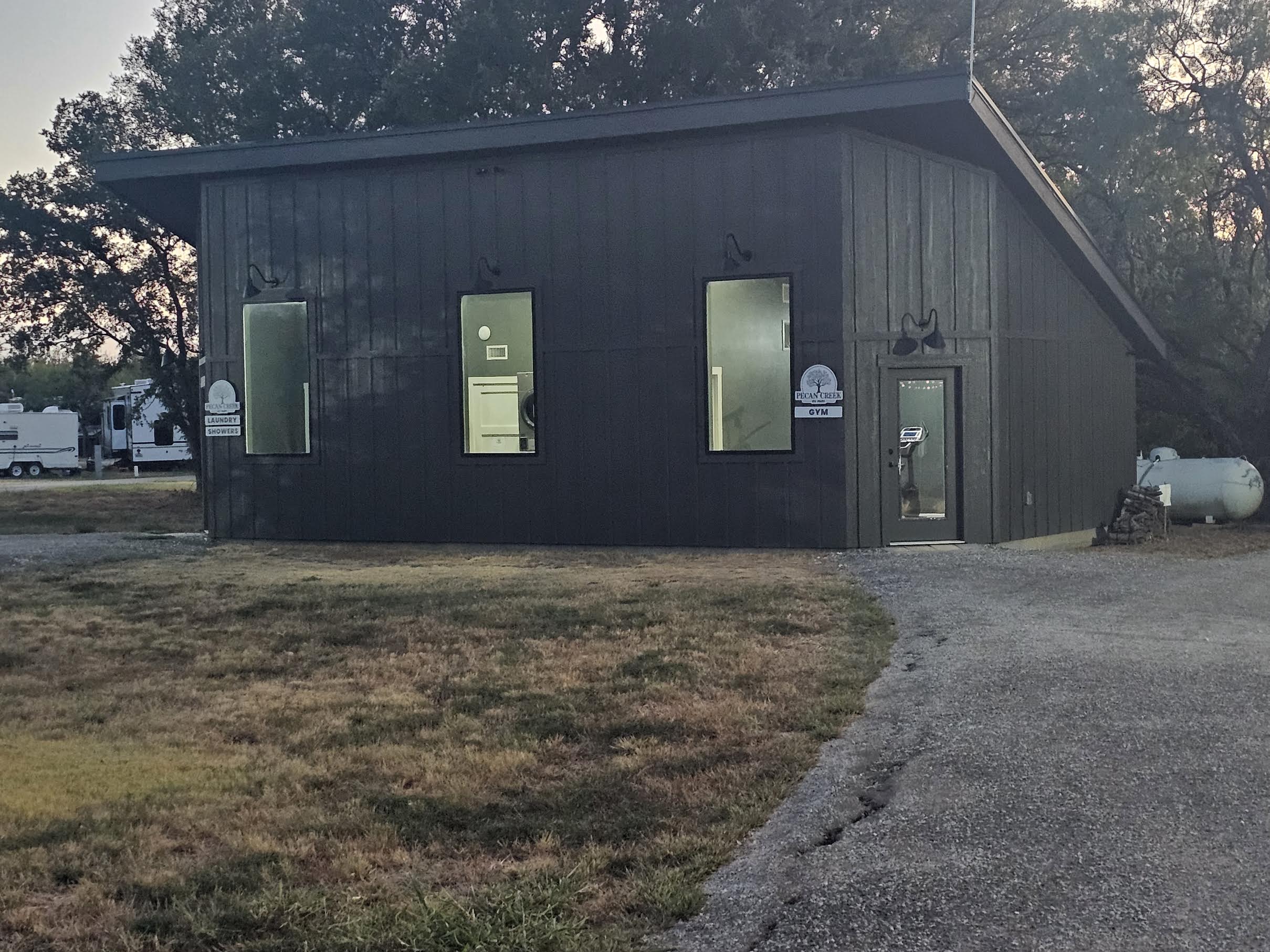 A dark-paneled building with three tall windows, a lit glass door, and outdoor lights. A curved driveway and dry grass are in the foreground. Trees and RVs are visible in the background.