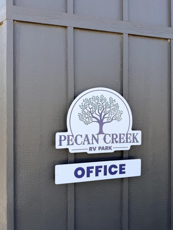 A sign on a dark wooden wall displays Pecan Creek RV Park with a tree logo above a smaller sign that says Office..